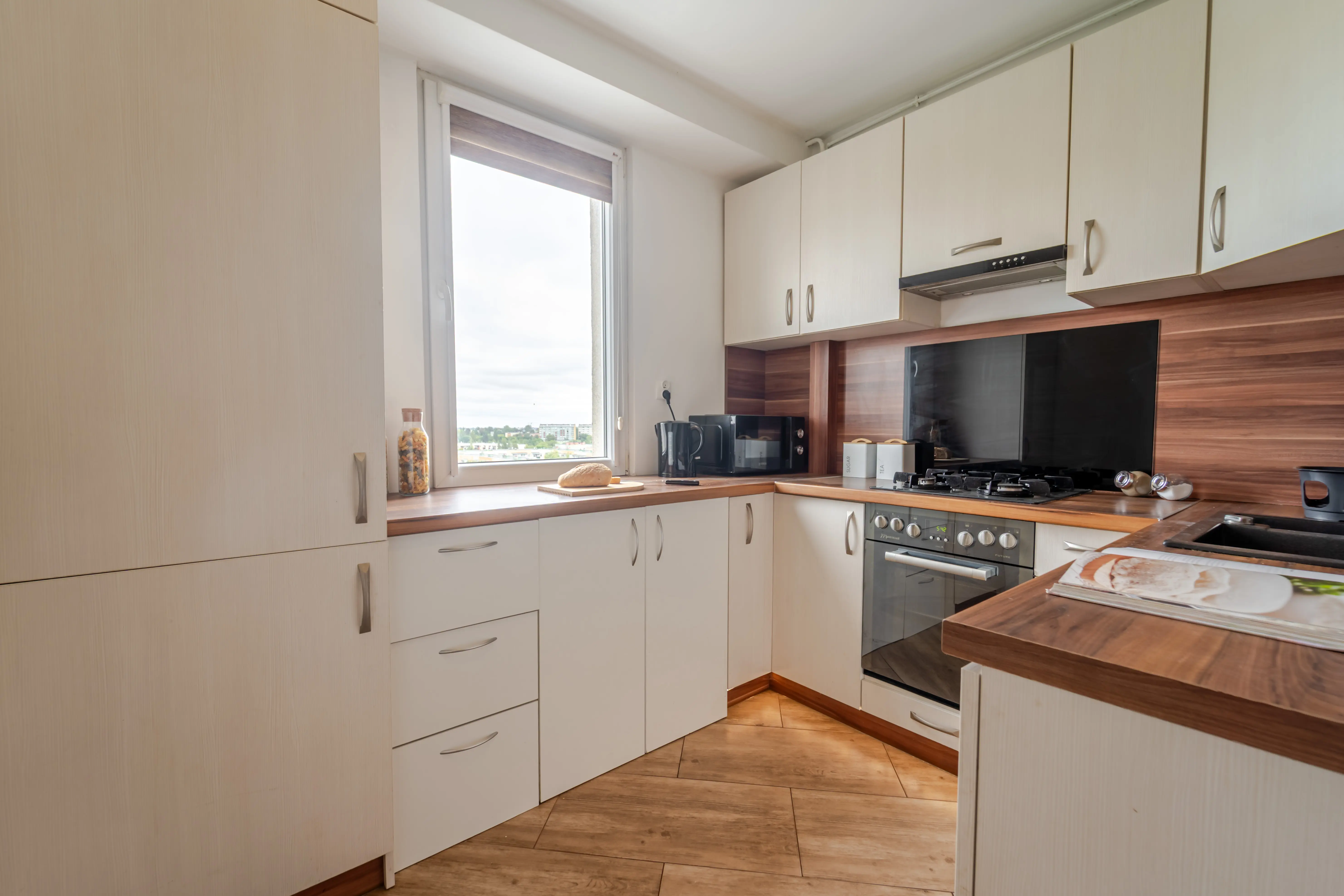 Klasyczna kuchnia w zabudowie narożnej z jasnymi frontami, drewnianym blatem i oknem zapewniającym naturalne oświetlenie blatu roboczego.