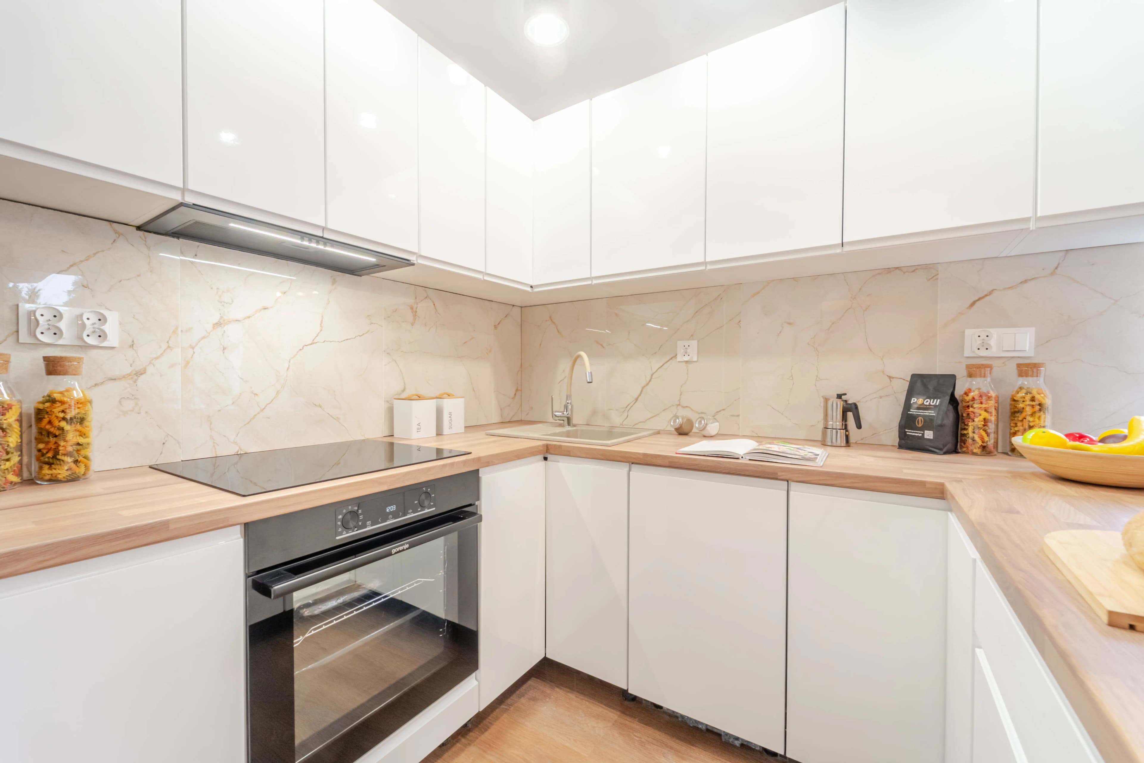 Jasna kuchnia w kształcie litery L z lakierowanymi szafkami sięgającymi sufitu i ścianą wykończoną płytkami imitującymi marmur.