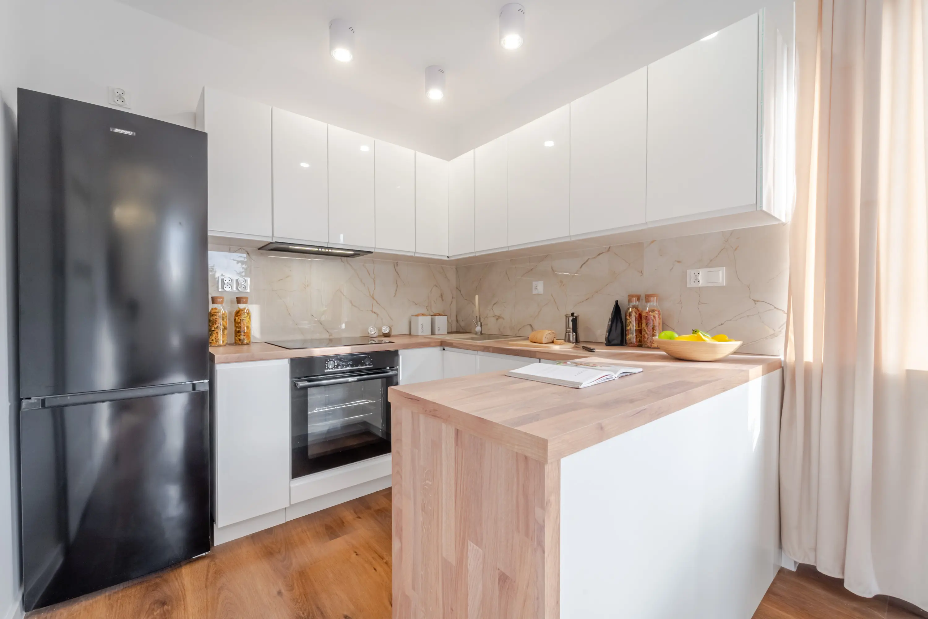 Nowoczesna biała kuchnia z drewnianym półwyspem, czarną lodówką wolnostojącą i oknem doświetlającym wnętrze.