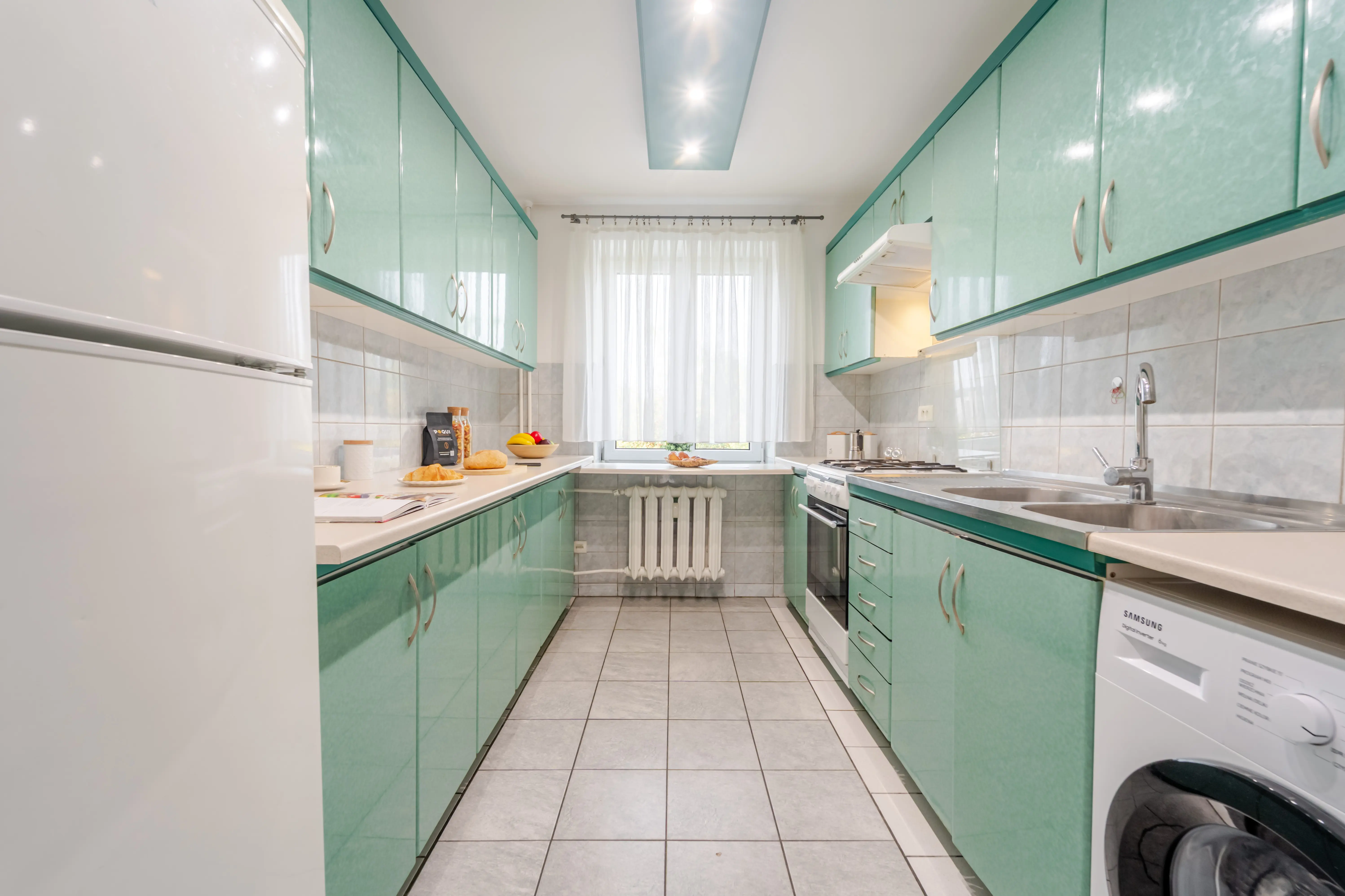 Dwustronna kuchnia w stylu retro z charakterystycznymi miętowymi frontami, oknem na końcu pomieszczenia i pralką w zabudowie.