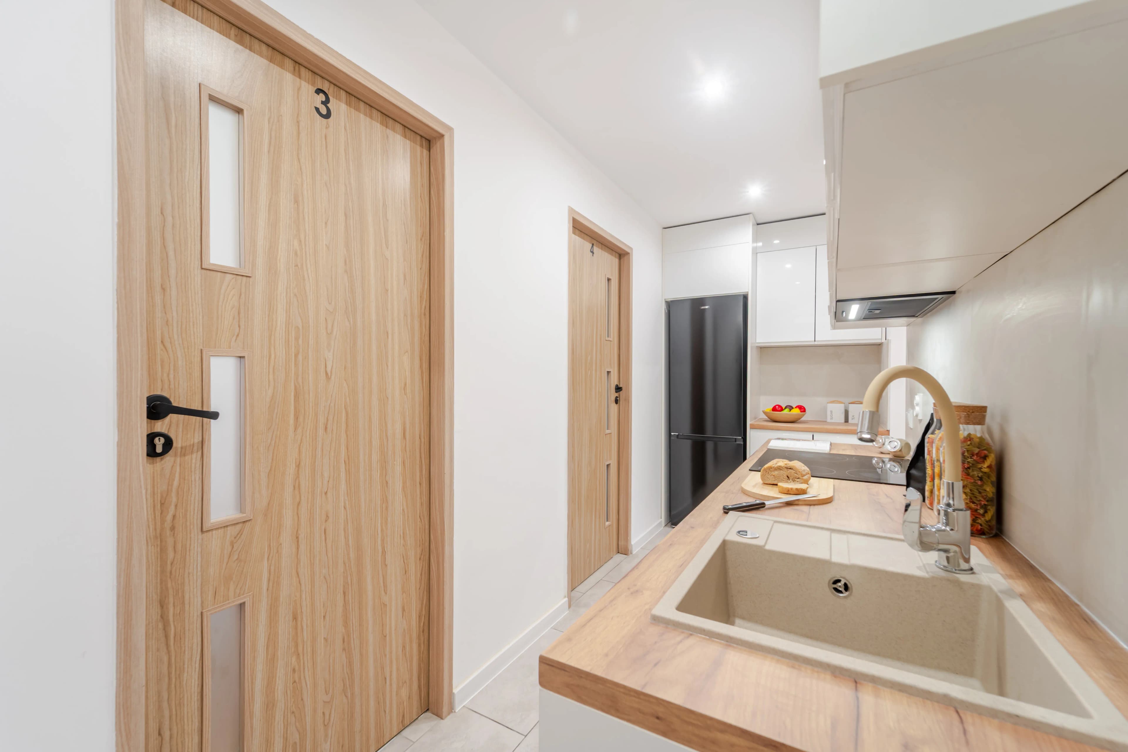 Nowoczesny aneks kuchenny w ciągu komunikacyjnym z granitowym zlewem, czarną lodówką i jasnymi drzwiami z okleiną drewnianą.