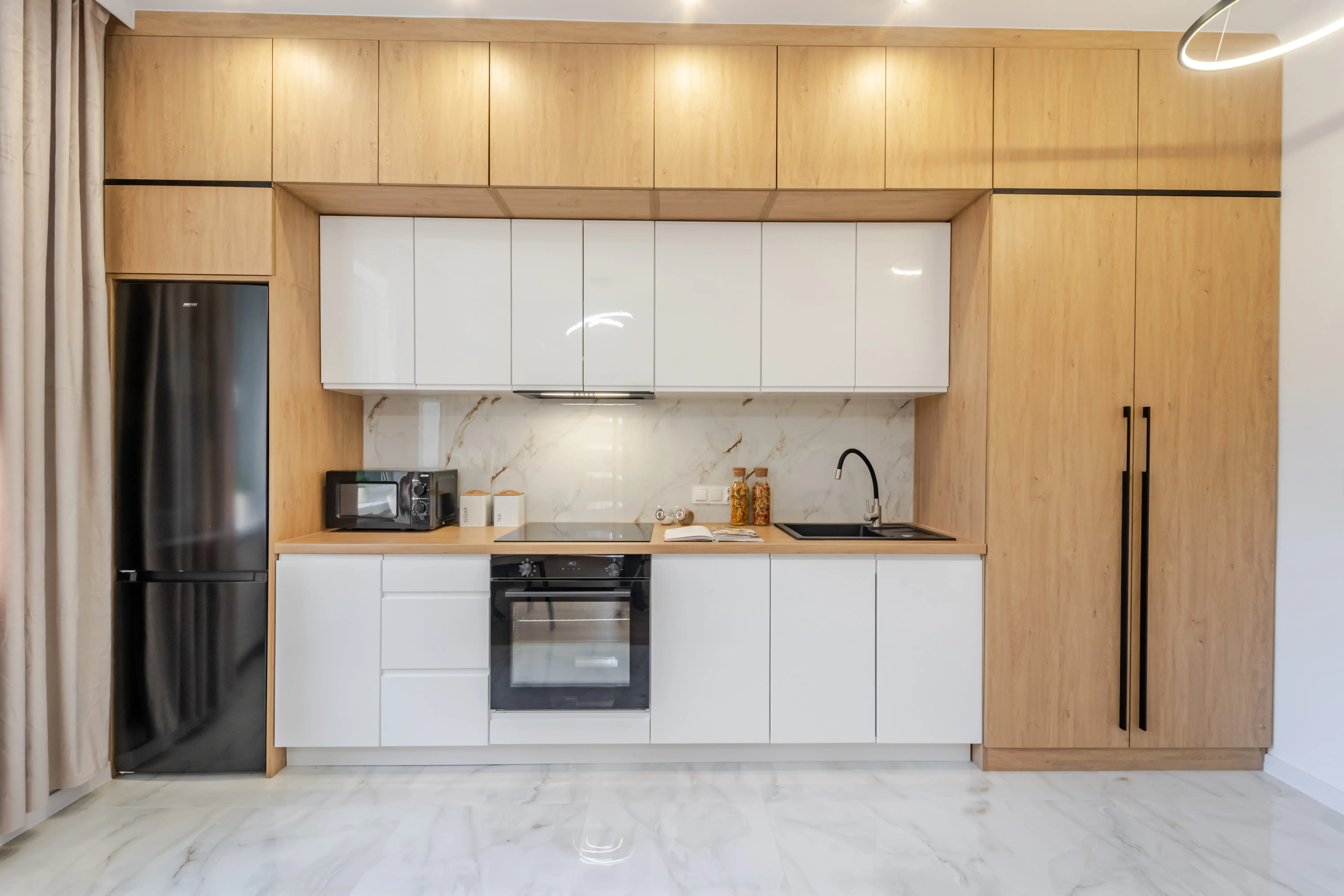 Symetryczna zabudowa kuchenna łącząca białe fronty z górnymi szafkami w odcieniu jasnego drewna i kontrastującą, czarną lodówką