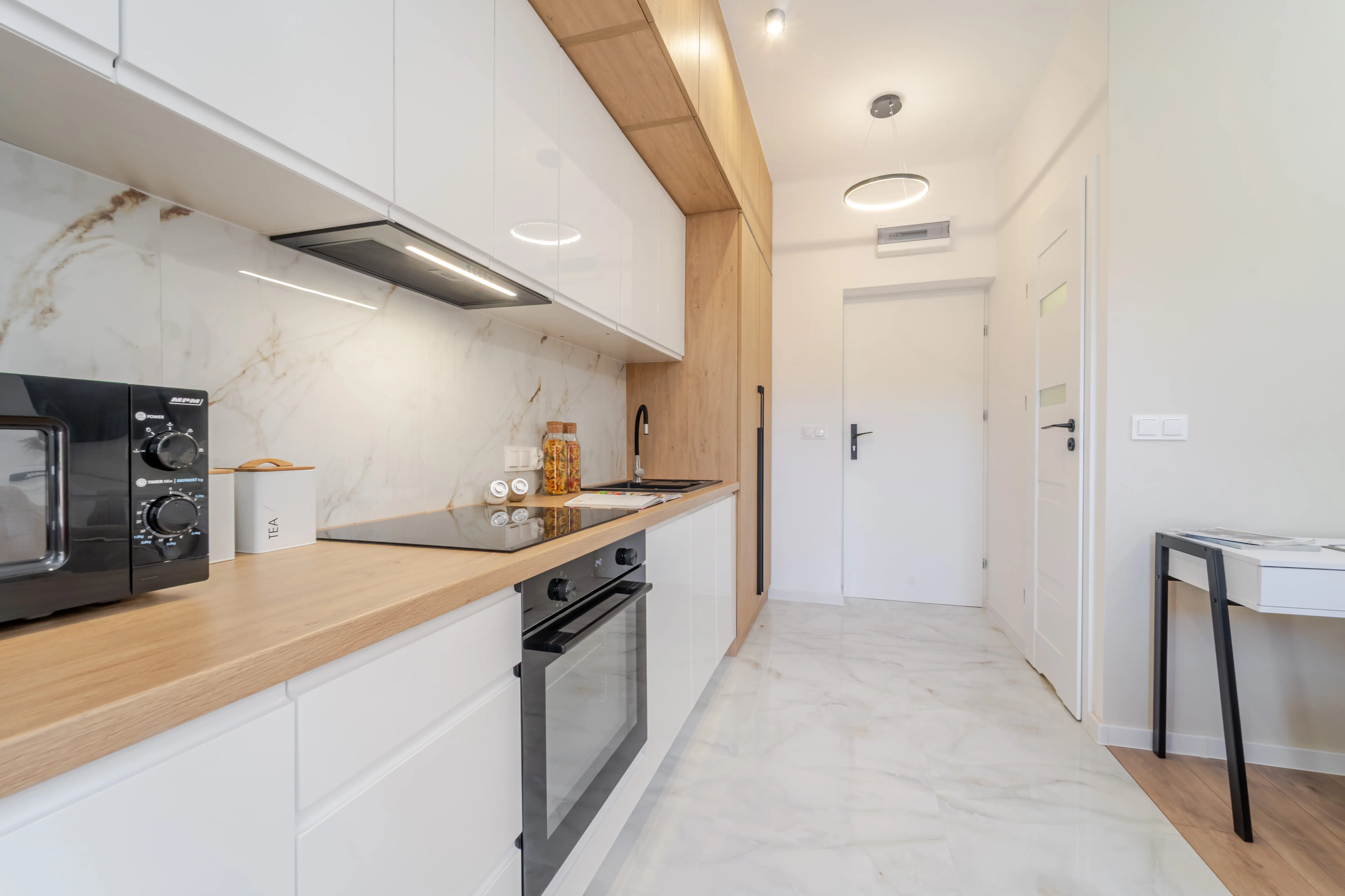 Widok wzdłuż nowoczesnej kuchni z marmurowym panelem ściennym, prowadzący wzrok ku jasnemu korytarzowi.