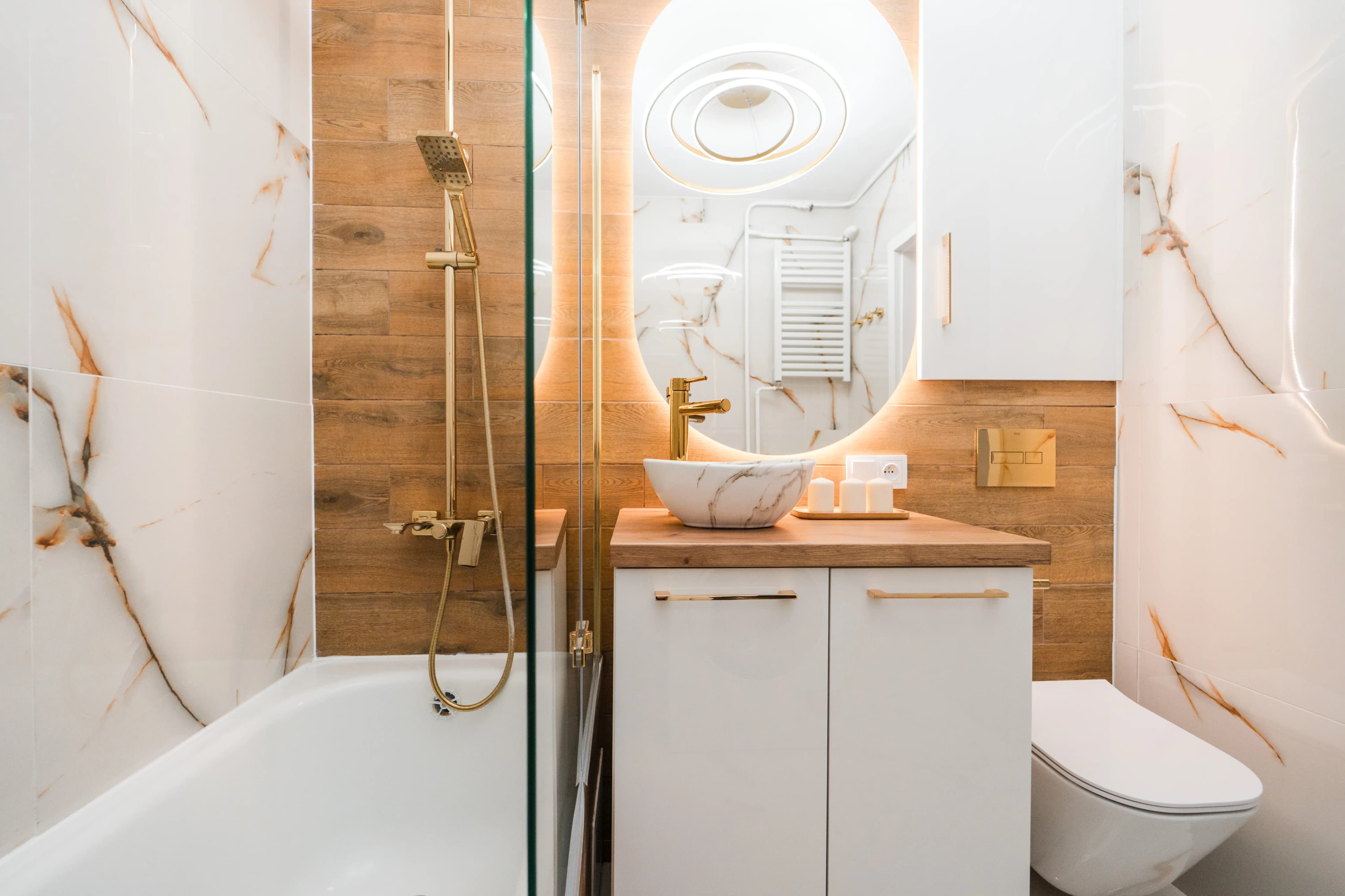 Elegancka łazienka wykończona marmurowymi płytkami i drewnem, wyposażona w złote armatury oraz podświetlane lustro owalne.