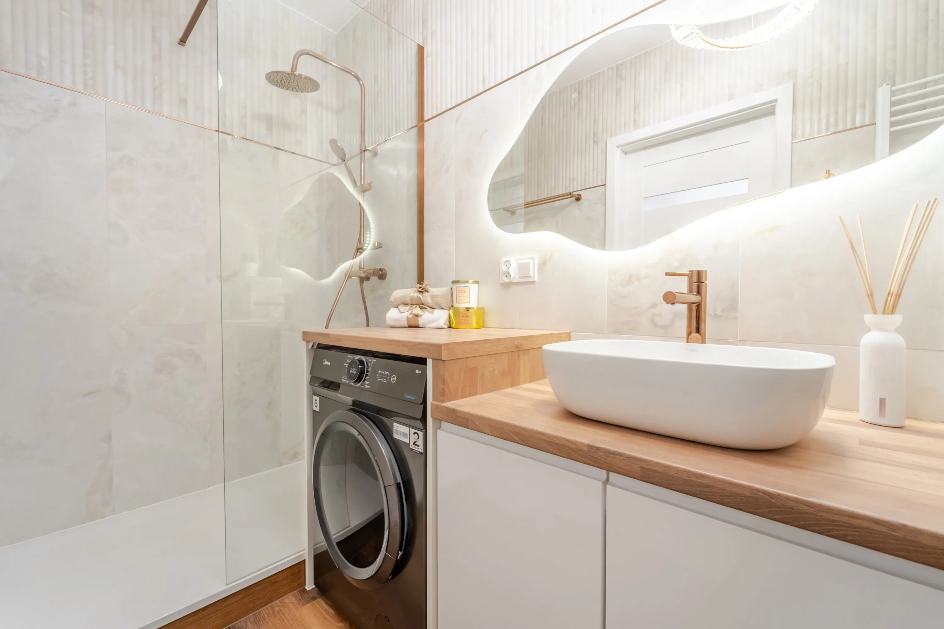 Elegancka strefa umywalkowa z drewnianym blatem, pod którym ukryto pralkę, dopełniona złotą armaturą i dekoracyjnym lustrem.