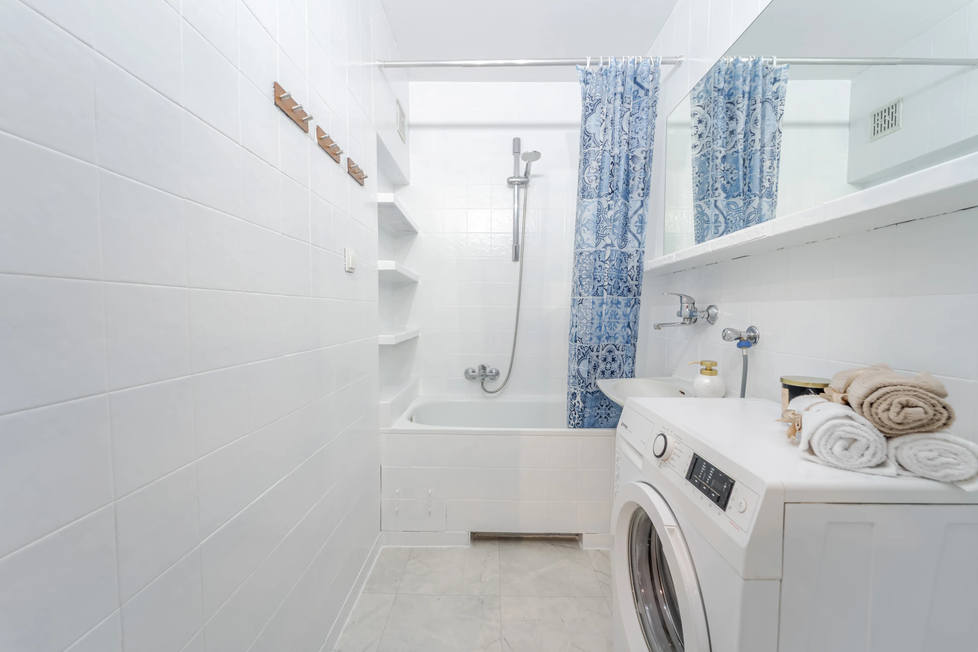 Jasna, klasycznie urządzona łazienka z białymi kafelkami, wanną oraz miejscem na pralkę.