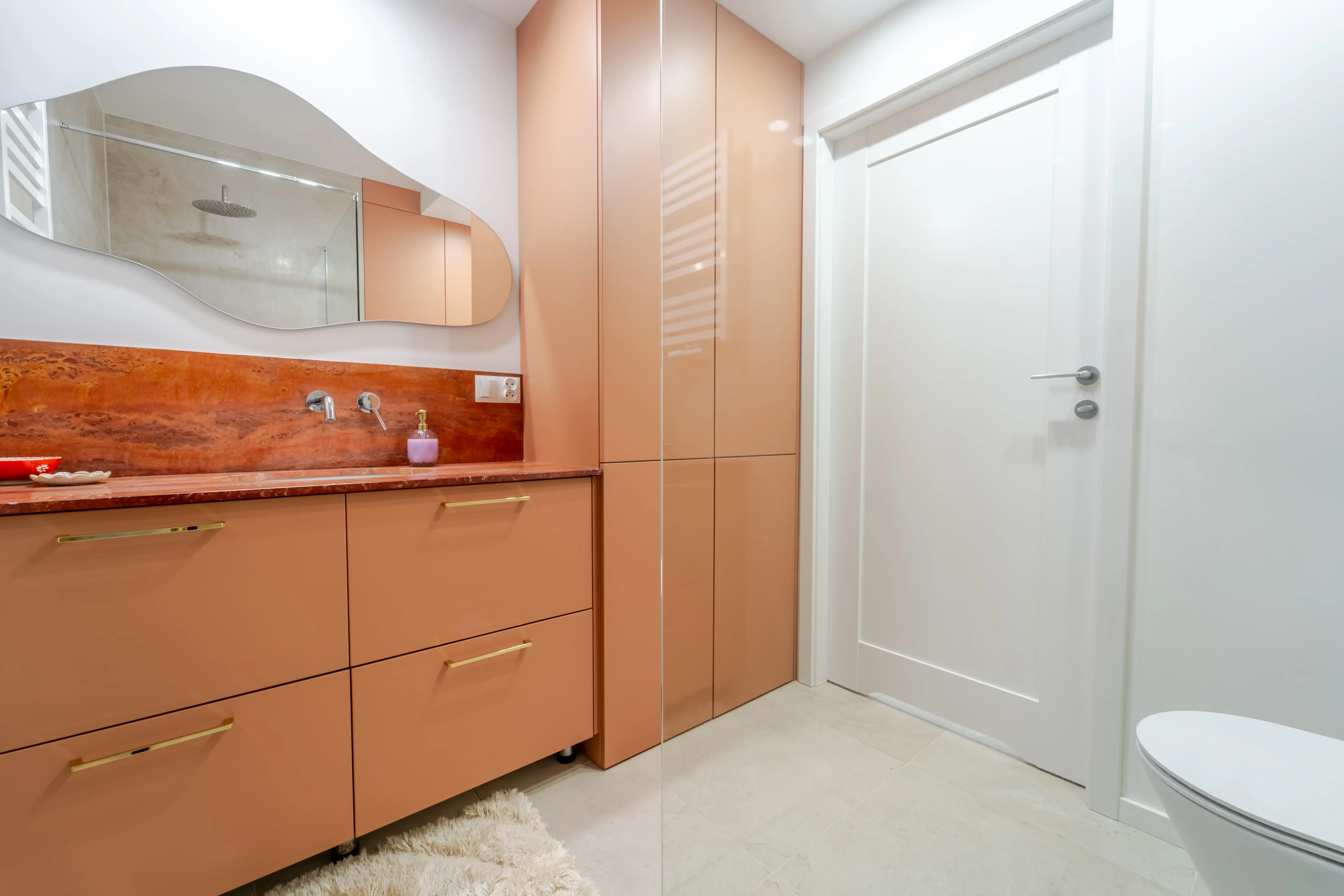 Odważna aranżacja łazienki z zabudową meblową w kolorze terakoty, kamiennym blatem i lustrem o kształcie chmury.