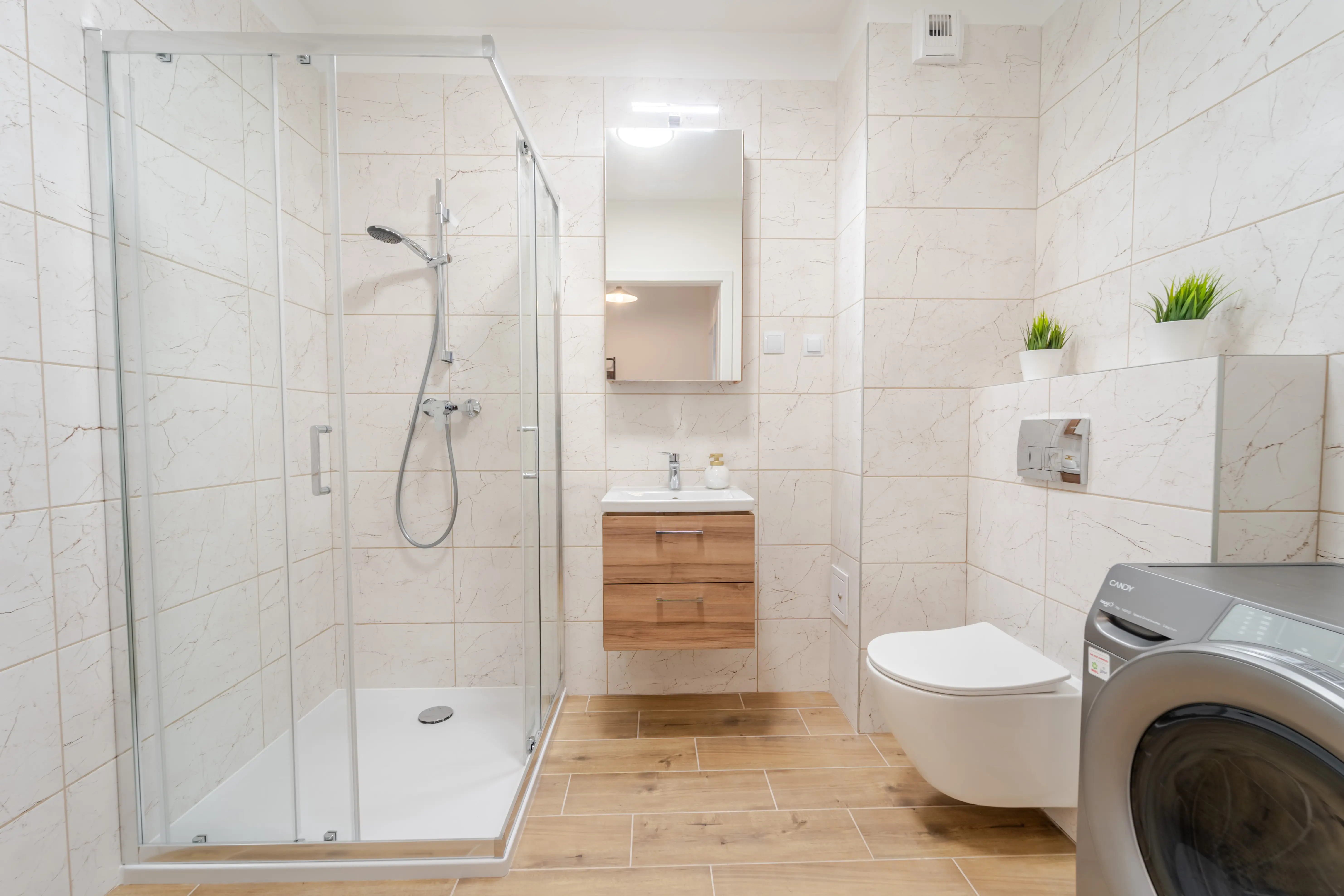 Jasna, funkcjonalna łazienka z narożną kabiną prysznicową, wiszącą szafką pod umywalkę i srebrną pralką.