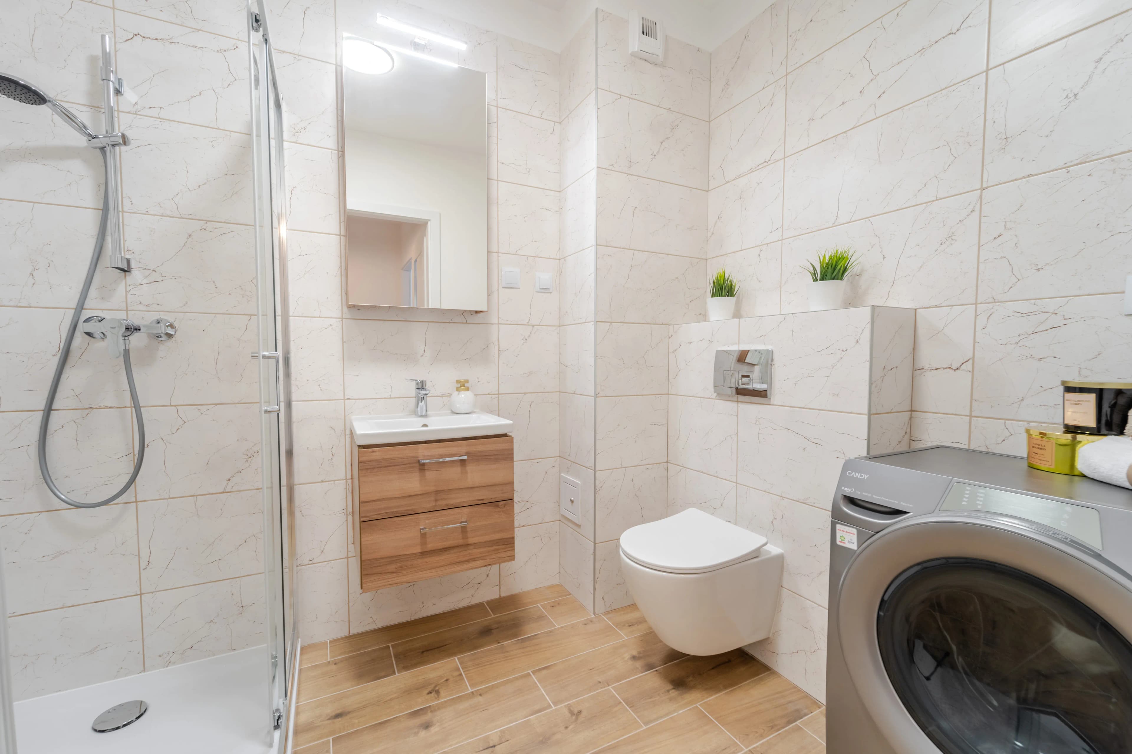 Jasna łazienka wykończona płytkami imitującymi marmur i drewno, wyposażona w szklaną kabinę prysznicową oraz wiszącą szafkę umywalkową.