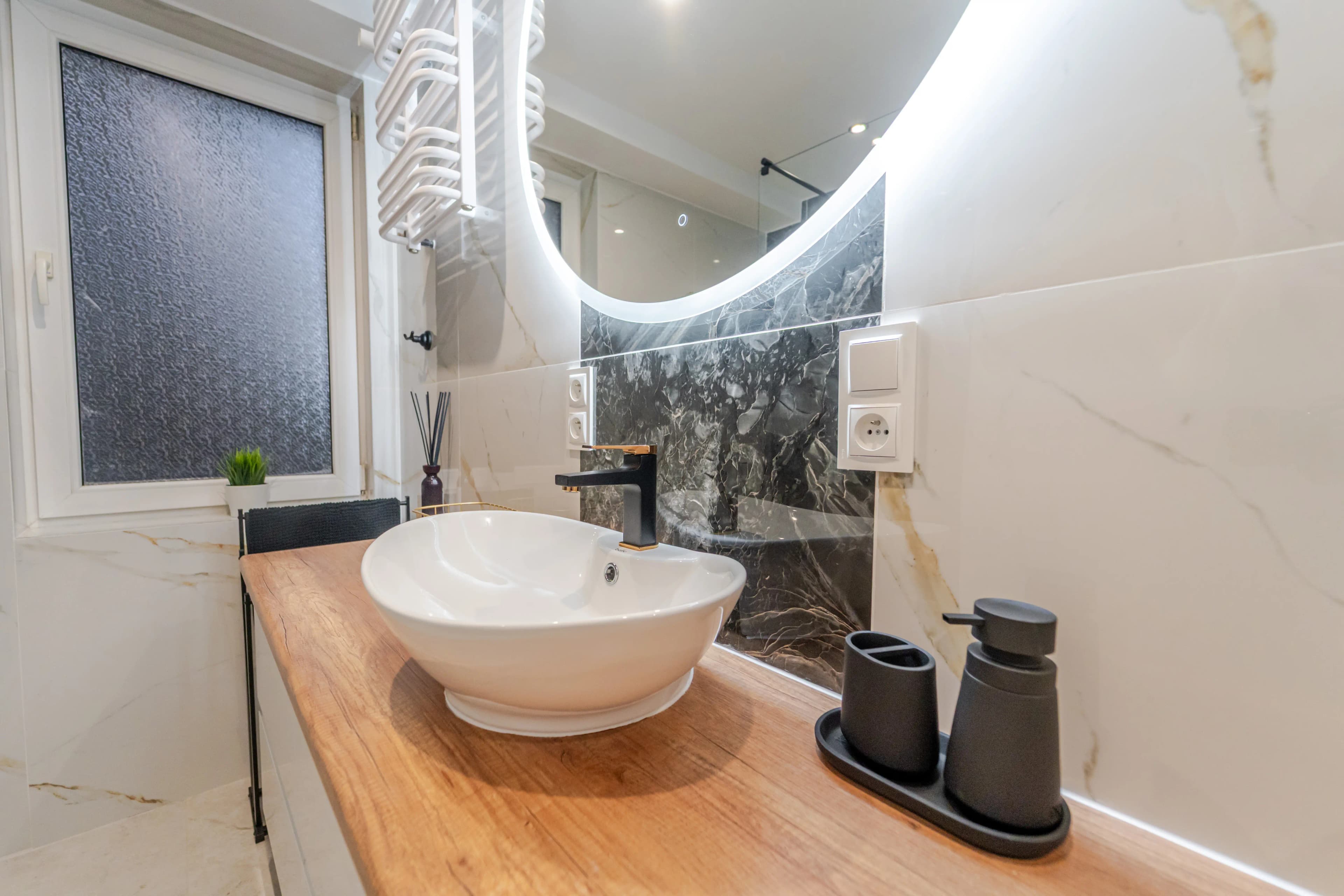 Zbliżenie na nablatową umywalkę w kształcie misy, czarną baterię ze złotym akcentem i podświetlane, okrągłe lustro.