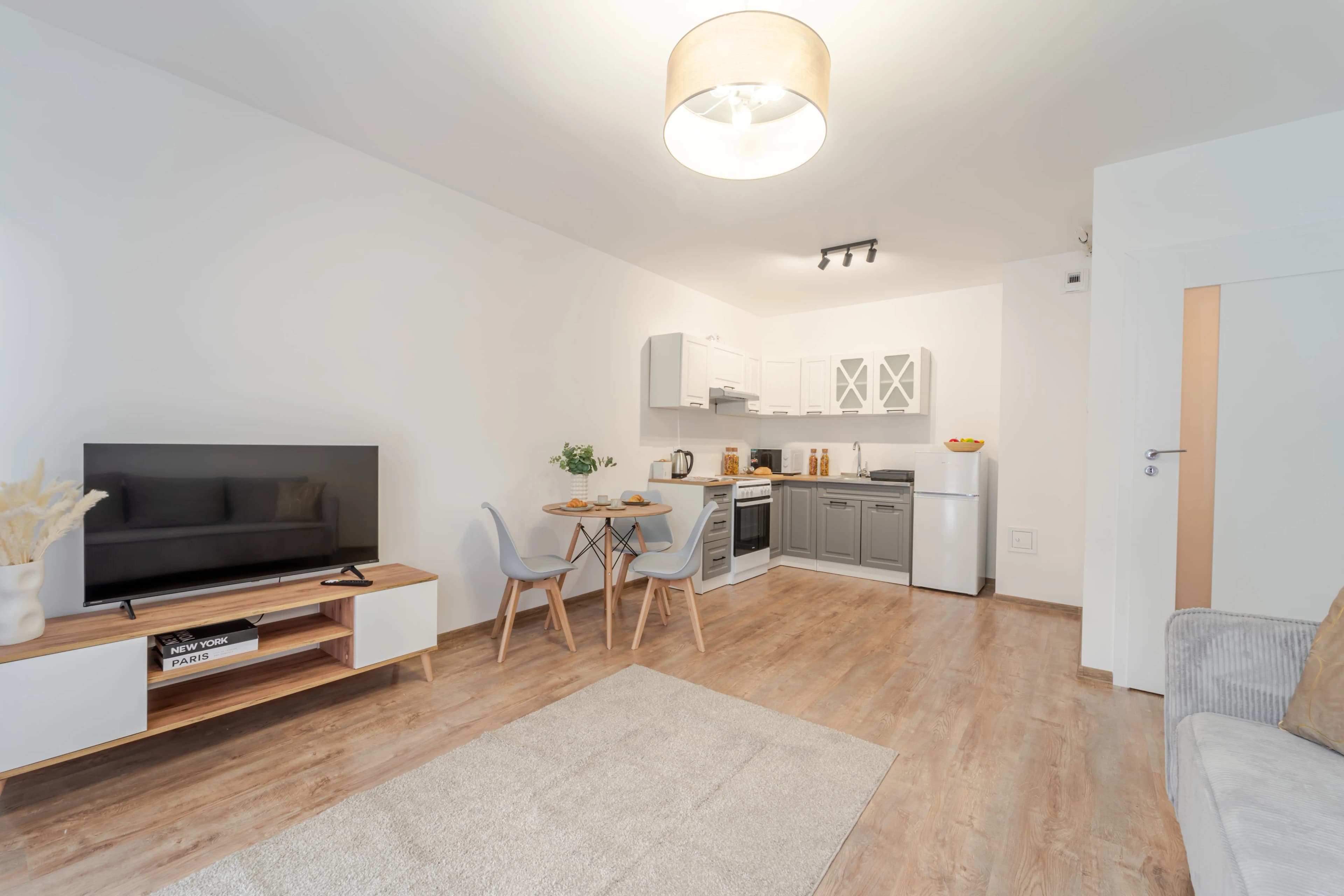 Minimalistycznie urządzony pokój dzienny z wygodną kanapą, roślinnością i otwartą kuchnią w tle.