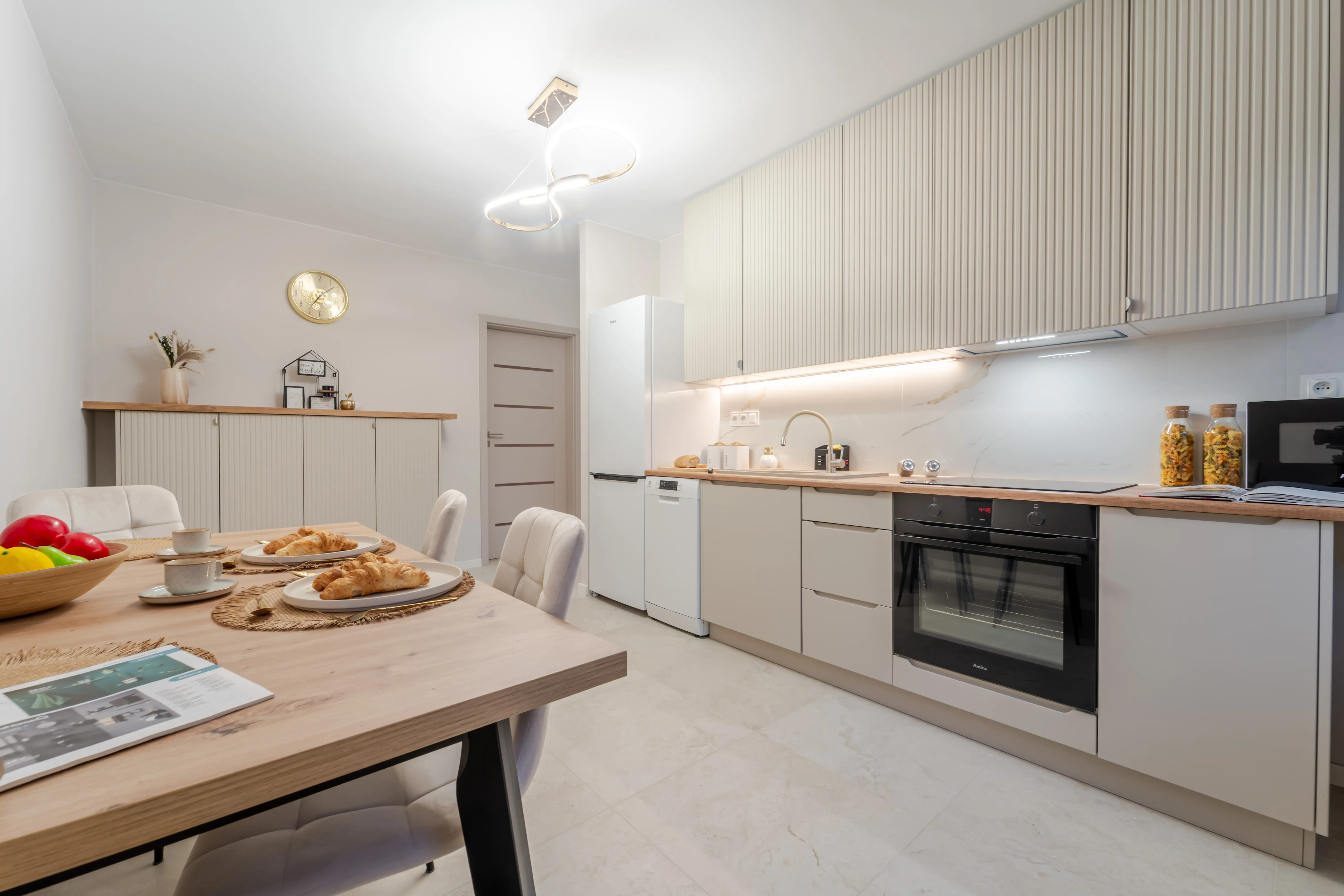 Funkcjonalna kawalerka łącząca strefę wypoczynkową z szarą sofą oraz aneks kuchenny z małym stołem jadalnianym.